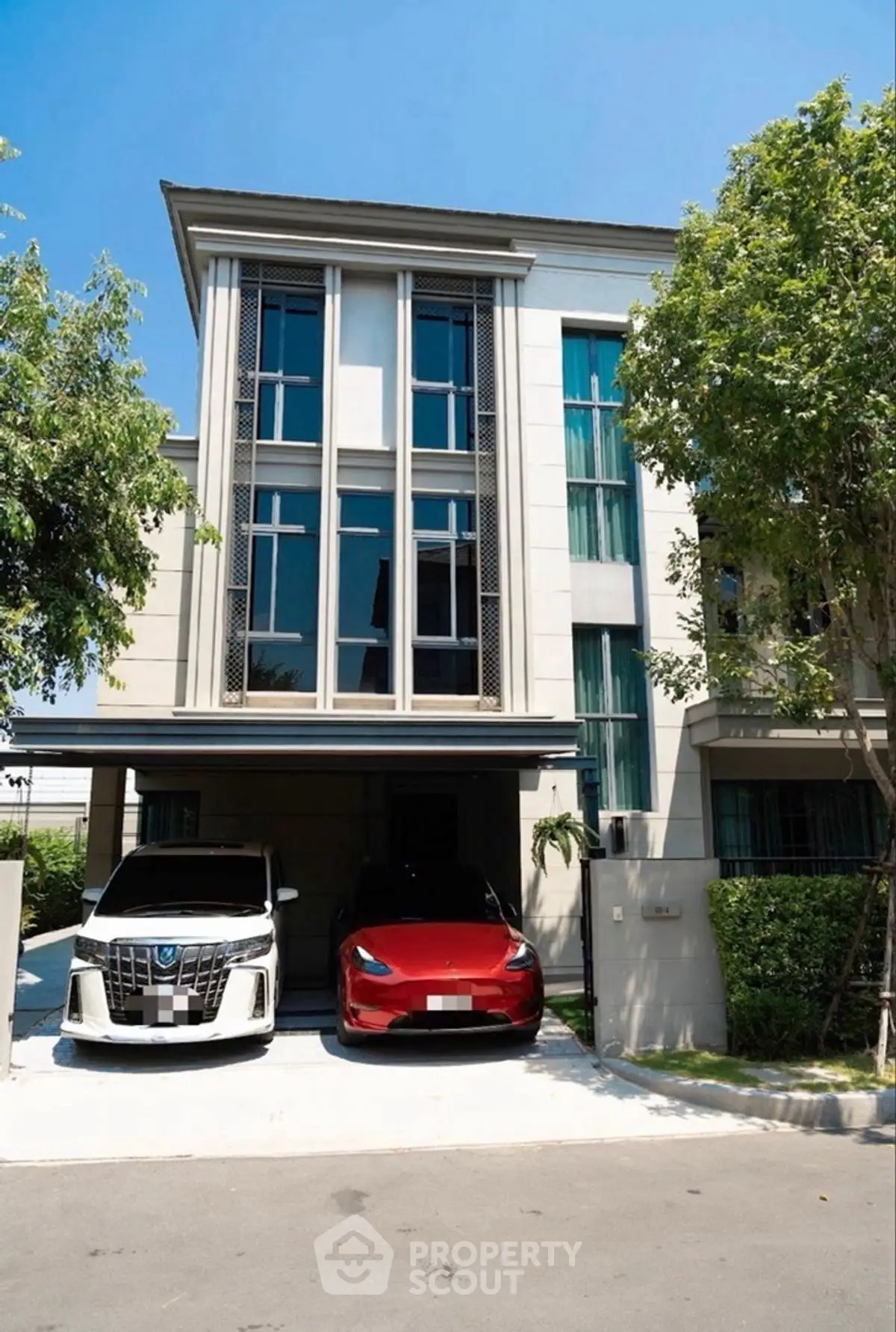 Modern three-story house with two cars parked in the driveway, surrounded by lush greenery.