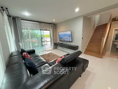 Modern living room with sleek black leather sofa and wall-mounted TV, featuring elegant staircase and large windows.