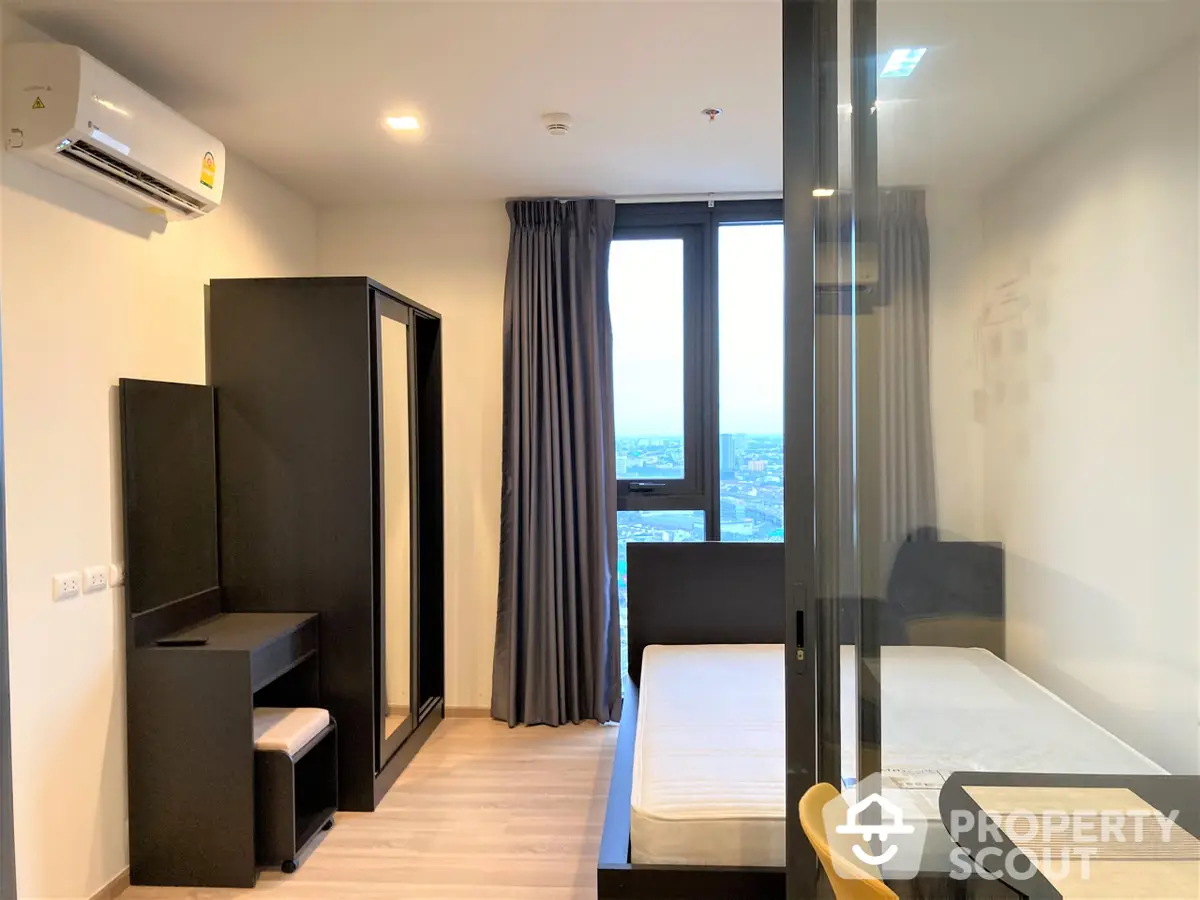 Modern studio apartment with sleek furniture, ample natural light, and a stunning city view through the large window, perfect for urban living.