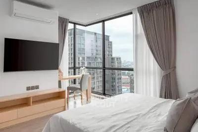 Modern bedroom with large windows offering stunning city views, featuring a sleek TV and cozy workspace.
