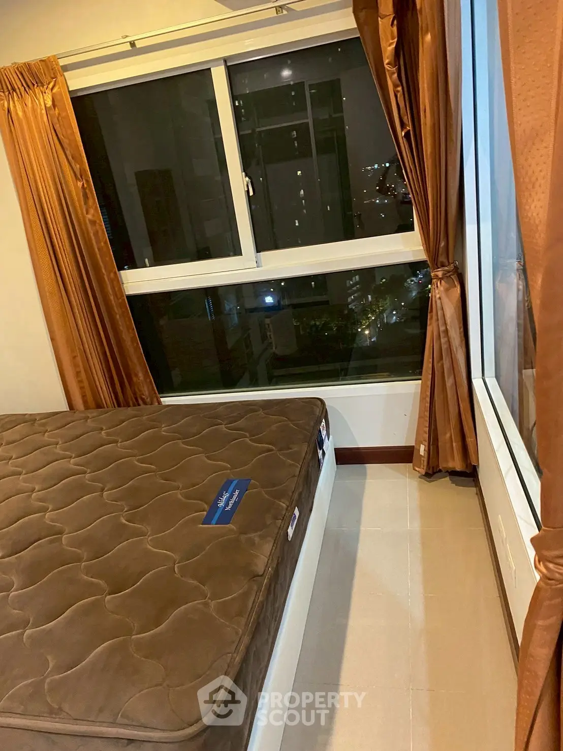 Cozy bedroom with large windows and city view, featuring elegant brown curtains and a comfortable mattress.