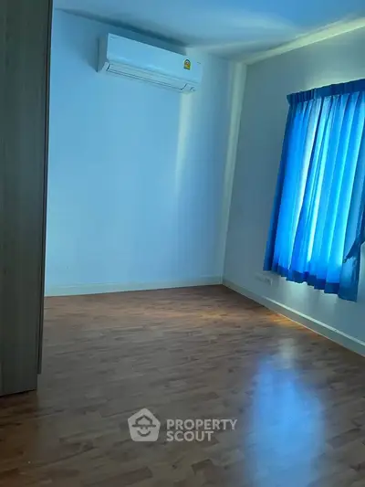 Spacious bedroom with wooden flooring and natural light from blue-curtained window, featuring air conditioning.