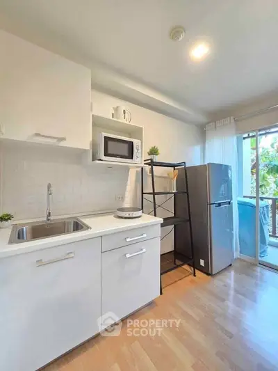 Modern kitchen with sleek white cabinets, microwave, and fridge, featuring natural light and wooden flooring.