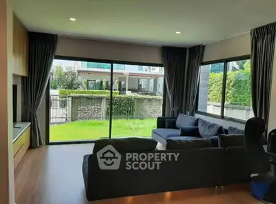 Spacious living room with large windows offering a lush garden view, featuring modern furniture and elegant curtains.