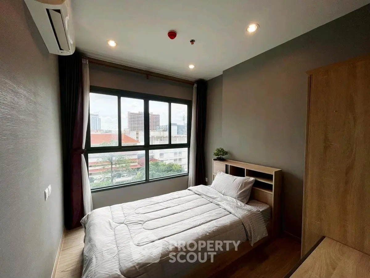 Cozy bedroom with large window and city view, featuring modern furnishings and natural light.