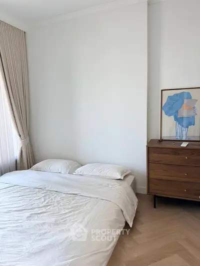 Cozy bedroom with minimalist decor and natural light, featuring a wooden dresser and elegant artwork.