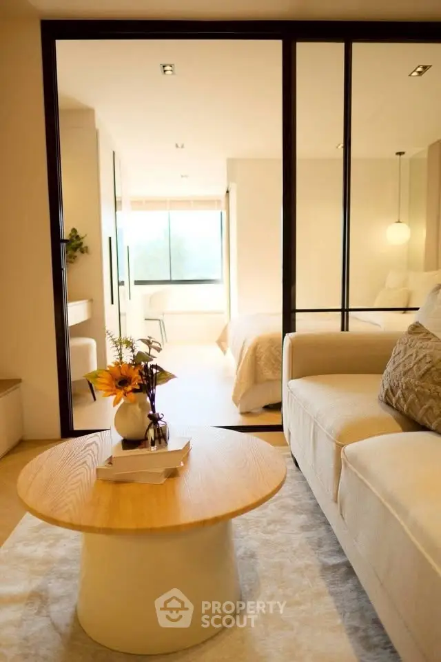 Modern living room with cozy sofa and stylish decor, featuring a view into a bright bedroom through glass doors.