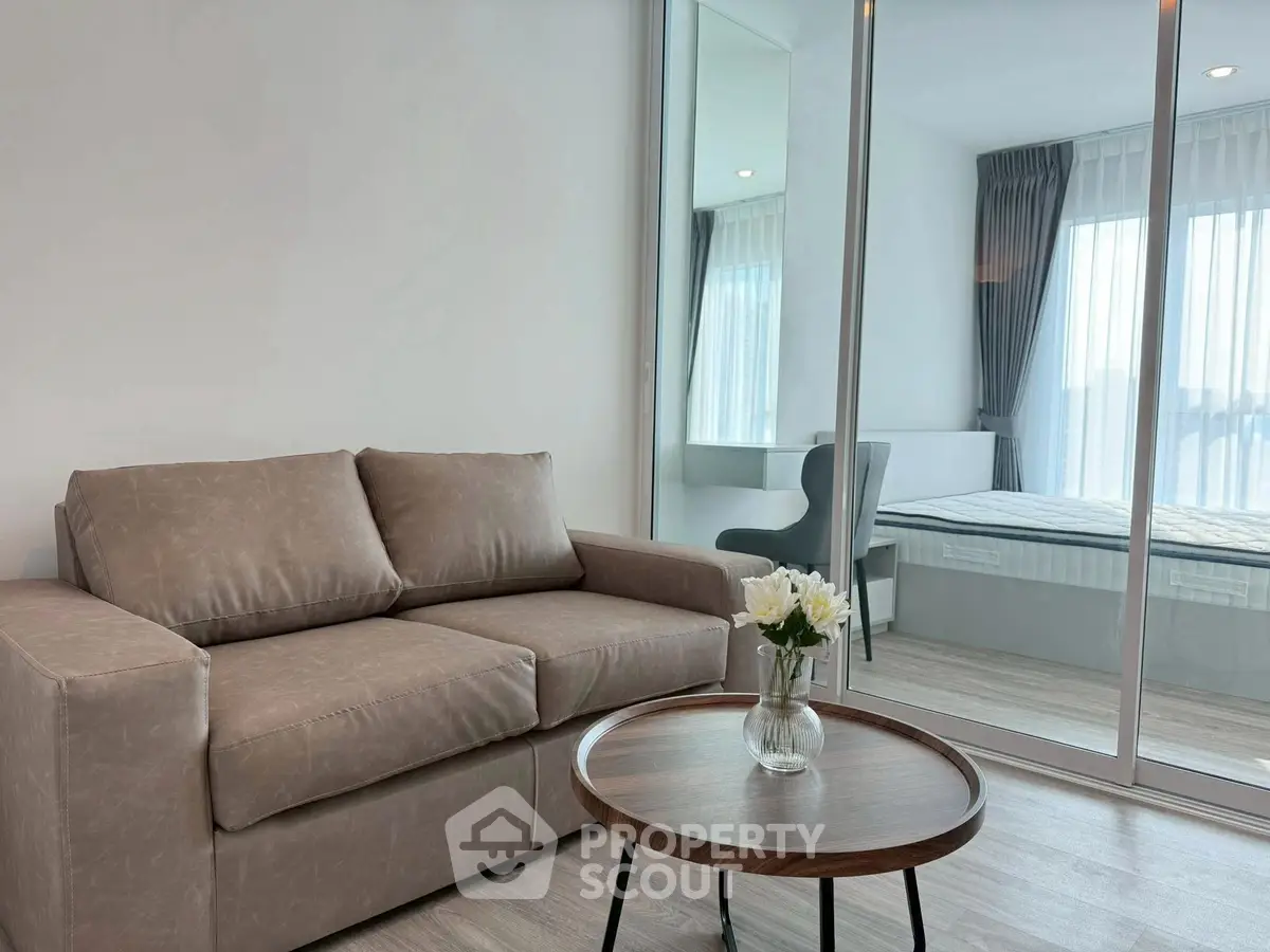 Modern living room with cozy sofa and glass partition leading to a bright bedroom.