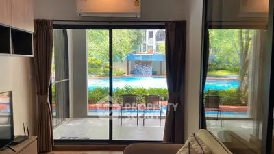 Modern living room with pool view through large glass doors, featuring cozy seating and lush greenery.