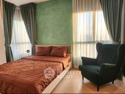 Cozy bedroom with elegant green accent wall and plush armchair, featuring natural light through large windows.