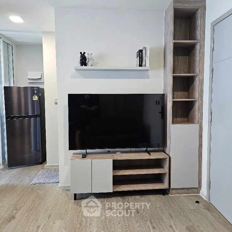 Modern living room with sleek TV unit and open shelving, adjacent to kitchen with stainless steel fridge.