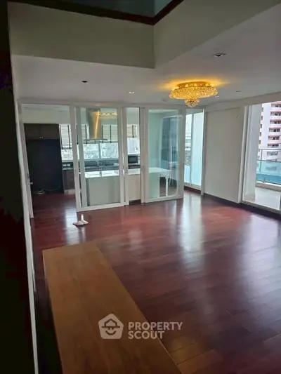 Spacious living room with hardwood floors and elegant chandelier, featuring a balcony with city views.