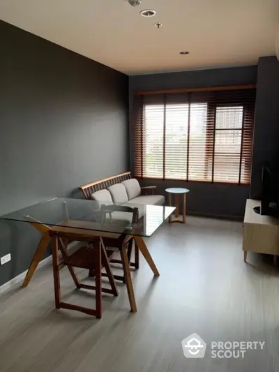 Modern living room with glass table and wooden furniture, large window with blinds.