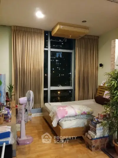 Cozy bedroom with wooden flooring, large window offering city views at night, and elegant drapery, perfect for urban living.