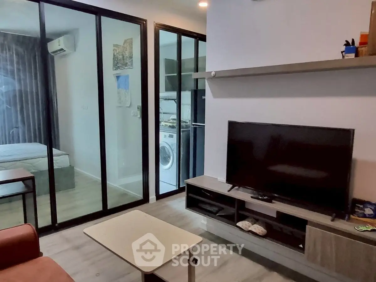 Modern living room with glass partition, TV unit, and view into bedroom and laundry area.