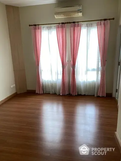 Spacious and well-lit empty room with polished wooden flooring, elegant pink curtains, and a modern air conditioning unit, ready for personalization.