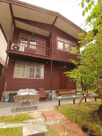 Charming two-story wooden house with lush garden and cozy outdoor seating area.