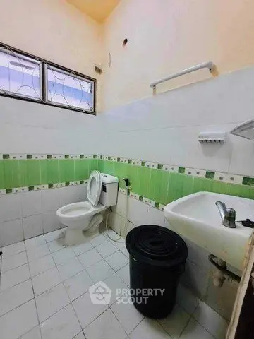 Compact bathroom with green and white tiles, featuring a toilet, sink, and window for natural light.