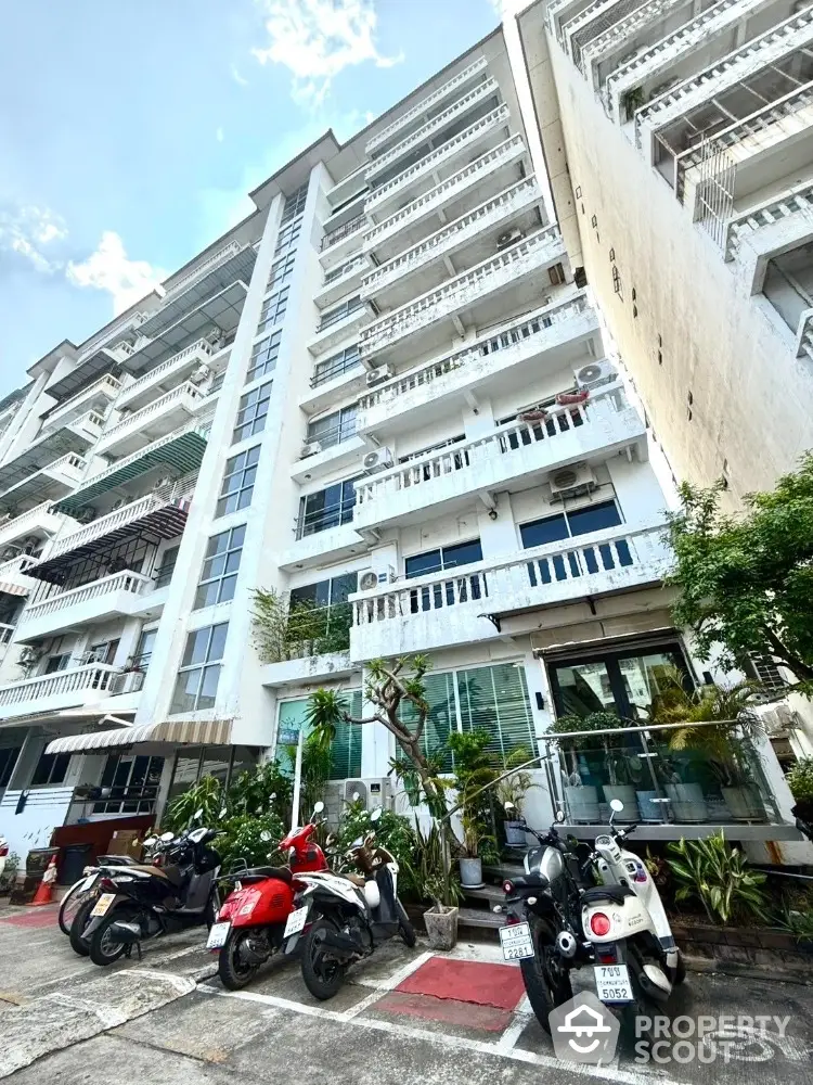 Modern apartment building with balconies and parking area featuring scooters, lush greenery enhances curb appeal.