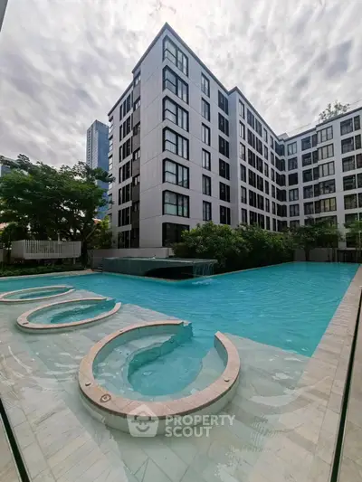 Modern apartment building with luxurious pool and water features under a cloudy sky.