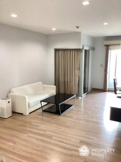 Spacious and well-lit living room with gleaming hardwood floors, modern white sofa, and sleek glass coffee table, leading to a sunlit balcony.