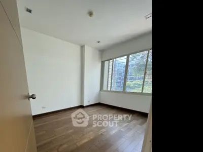 Spacious empty room with wooden flooring and large window offering natural light.