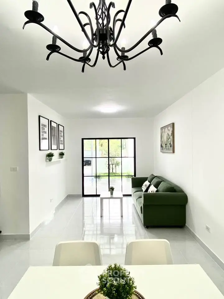 Spacious living room with modern chandelier and green sofa, bright natural light through large windows.