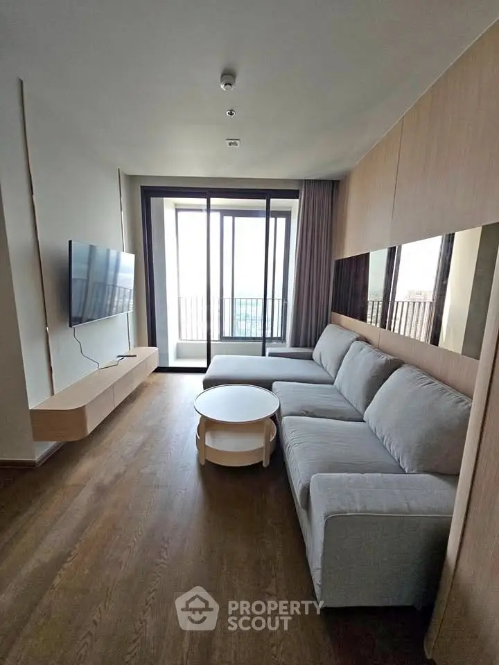 Modern living room with large sofa and wall-mounted TV, featuring floor-to-ceiling windows and wooden flooring.