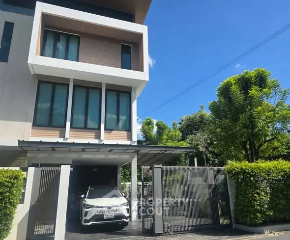 Modern three-story house with carport and lush greenery under a clear blue sky.