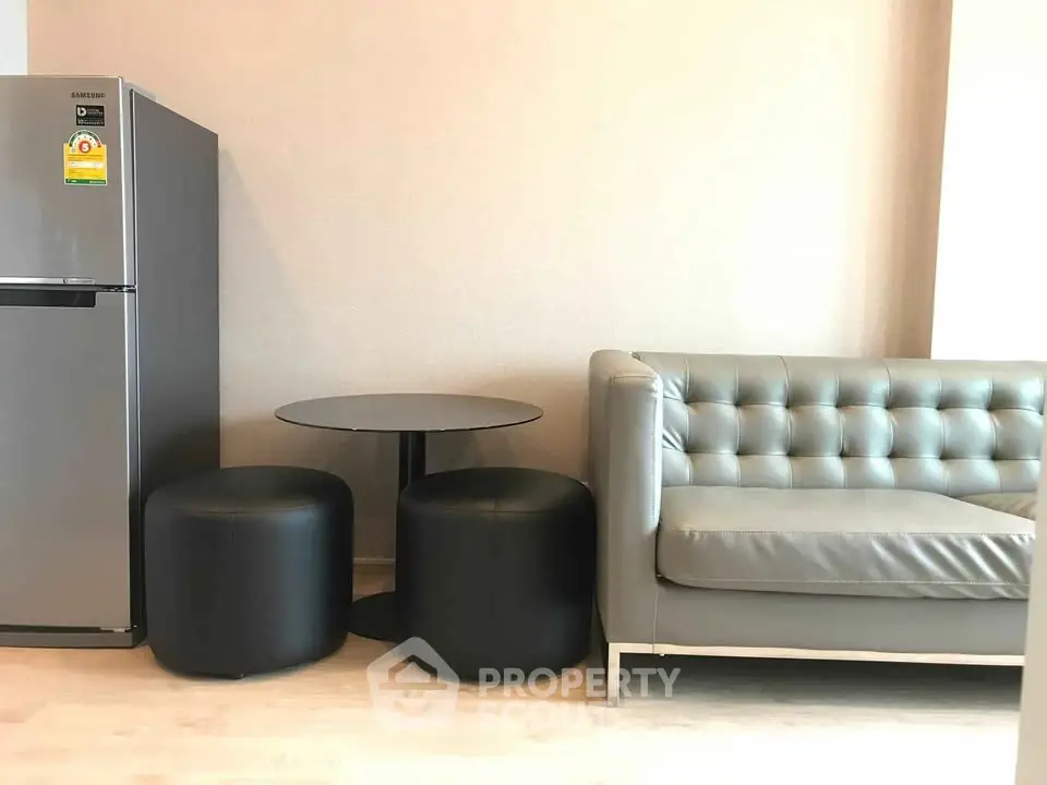 Modern living room with sleek gray sofa, compact dining set, and stainless steel fridge.
