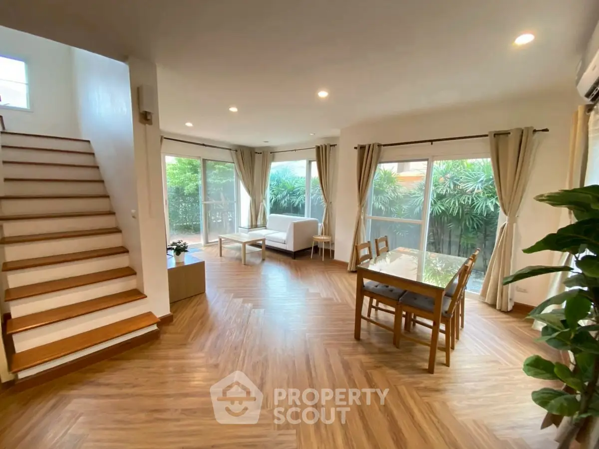 Spacious living room with wooden flooring and large windows overlooking greenery.