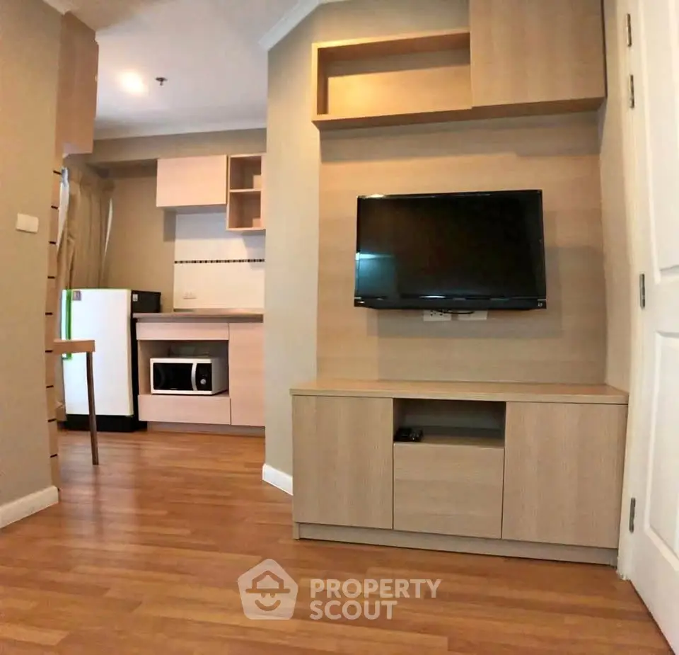 Modern apartment interior with wooden flooring, wall-mounted TV, and compact kitchen area.