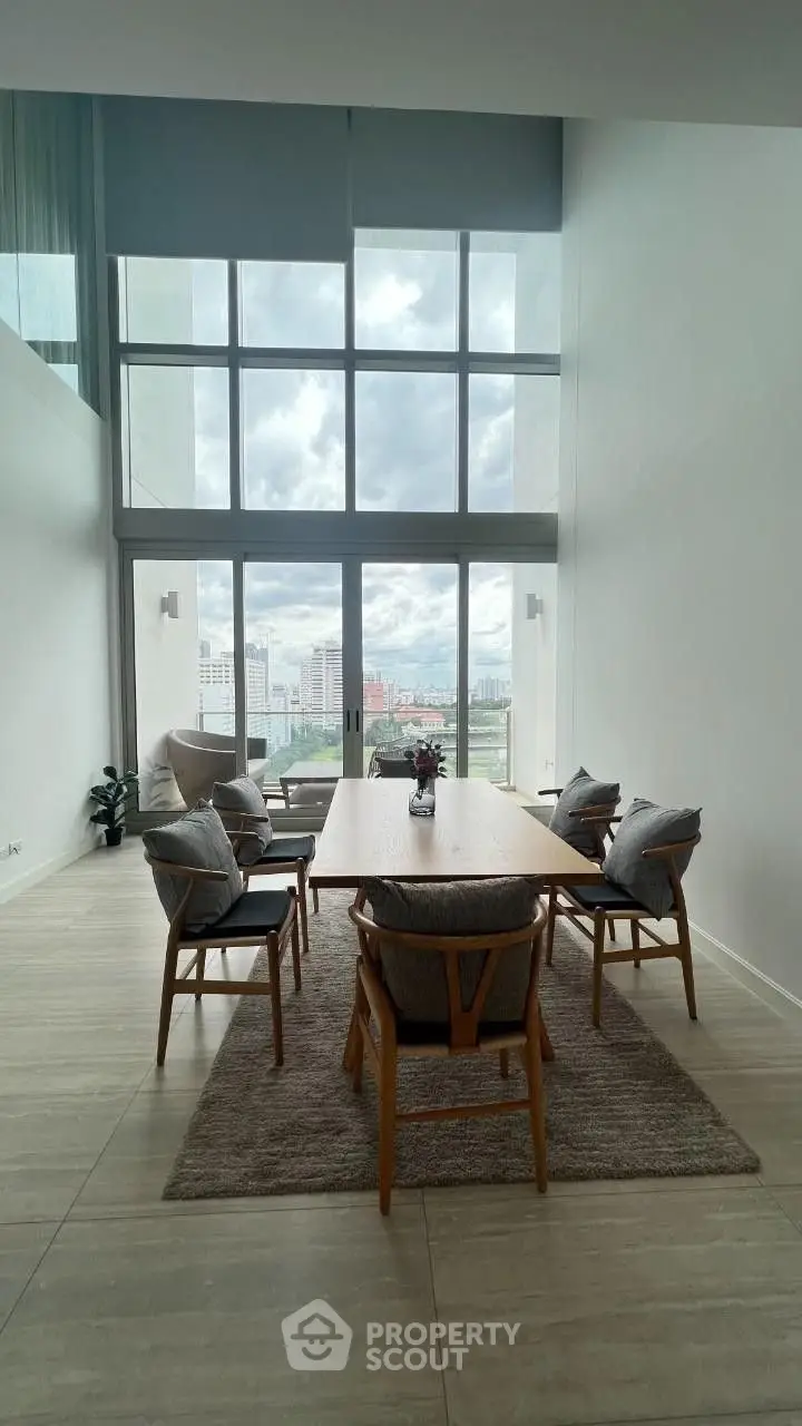 Spacious living room with high ceiling and large windows offering panoramic city views, complemented by a stylish dining set and cozy seating area.
