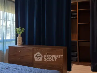 Elegant bedroom with wooden dresser and open walk-in closet, featuring dark curtains and natural light.