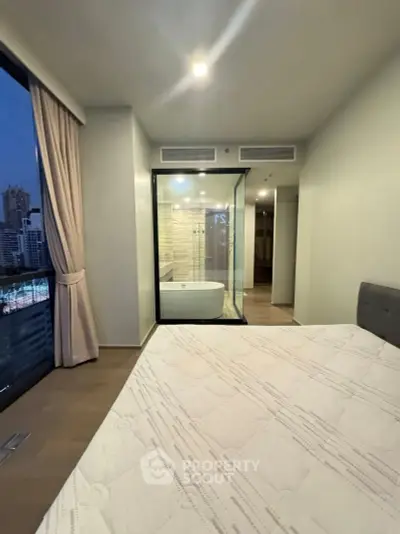 Modern bedroom with city view and glass-walled bathroom featuring a bathtub.