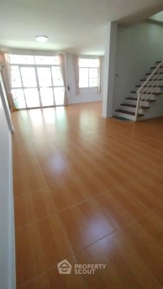 Spacious living room with wooden flooring and staircase, ample natural light from large windows.