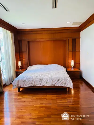 Elegant bedroom with polished hardwood flooring and rich wooden wall paneling, featuring a large bed with classic bedding, flanked by matching nightstands and lamps, and ample natural light from the window.