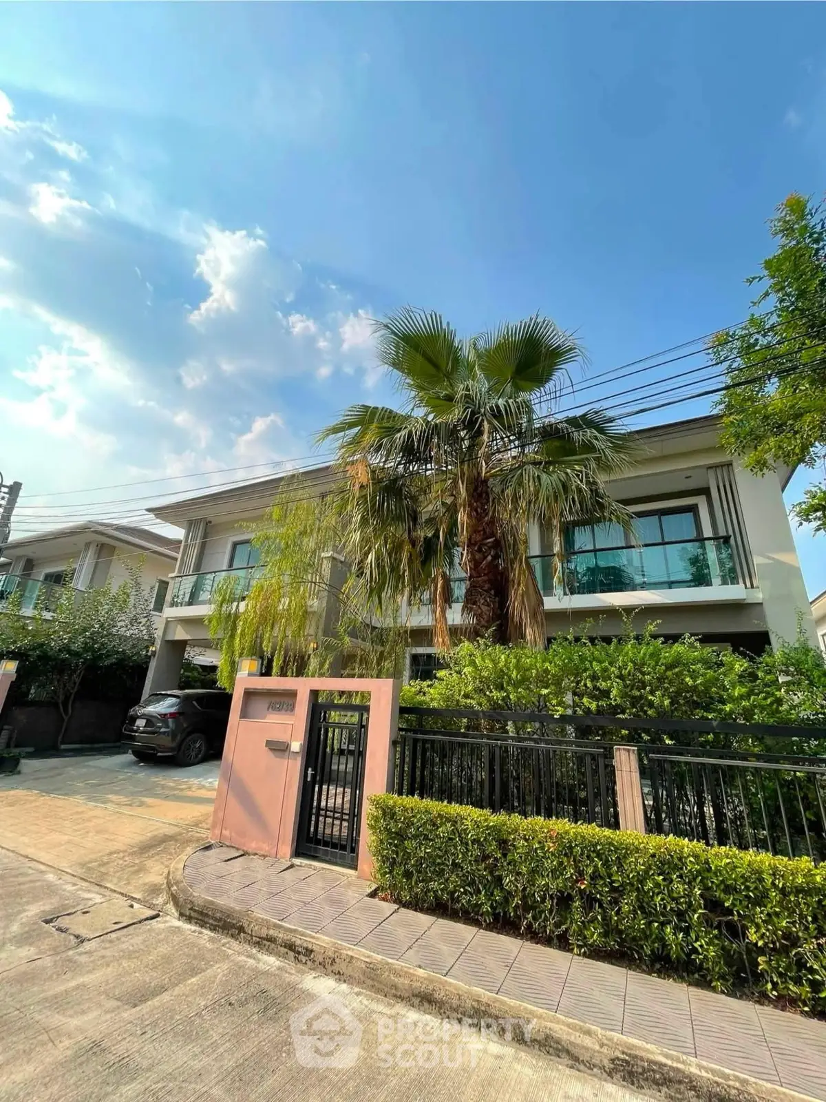 Modern two-story house with lush greenery and palm tree, featuring a gated entrance and driveway.