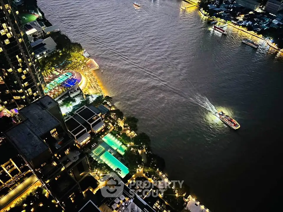 Stunning aerial view of riverside luxury apartments with illuminated pools and vibrant cityscape at night.