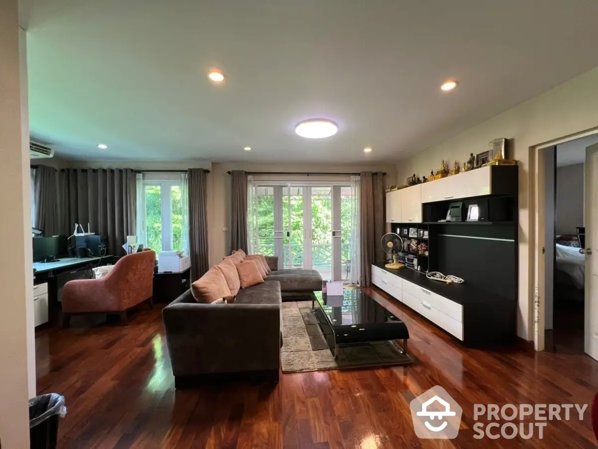 Spacious living room with polished hardwood floors, modern furniture, and ample natural light from large windows overlooking greenery.