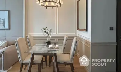 Elegant dining area with a modern glass table, plush chairs, and a chic chandelier, complemented by classic wall paneling and neutral tones.