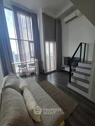 Modern loft-style living room with high ceilings and large windows, featuring a cozy sofa and sleek staircase.