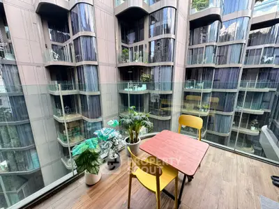 Modern balcony with glass railing overlooking contemporary building facade, featuring stylish outdoor seating and lush plants.