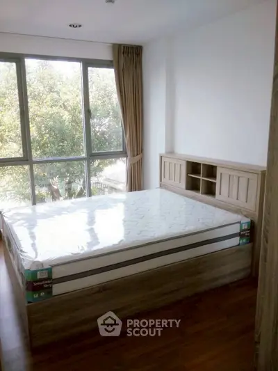 Cozy bedroom with large window and natural light, featuring a new mattress and wooden flooring.