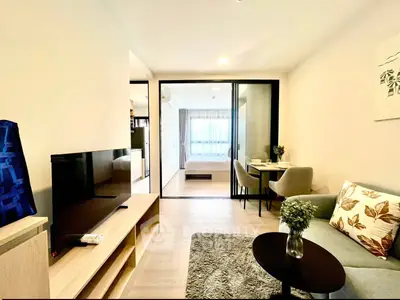 Modern living room with cozy seating and TV, leading to a bright bedroom through glass doors.