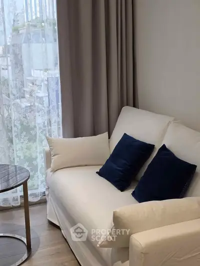 Cozy living room with elegant white sofa and blue cushions, featuring a round glass table and sheer curtains.