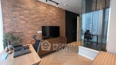 Chic urban living room with exposed brick wall, sleek entertainment unit, and herringbone wood flooring, leading to a modern glass-partitioned study.