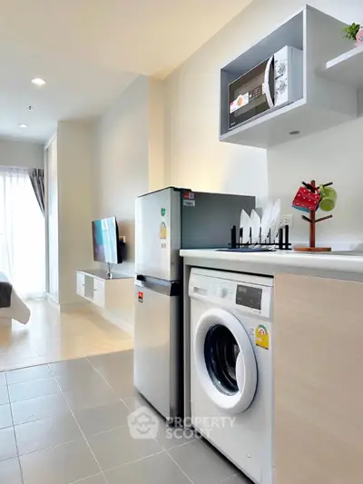 Modern apartment kitchen with washing machine, fridge, and microwave, featuring sleek design and natural light.
