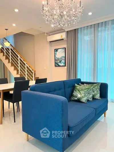 Modern living room with blue sofa, elegant chandelier, and dining area in open layout.