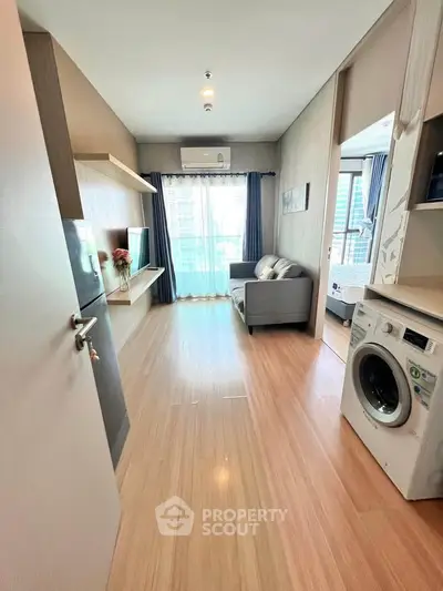 Modern apartment interior with washing machine and cozy living area, featuring large windows and wooden flooring.